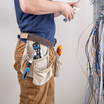 Electrician builder at work, examines the cable connection in the electrical line in the fuselage of an industrial switchboard. Professional in overalls with an electrician's tool.